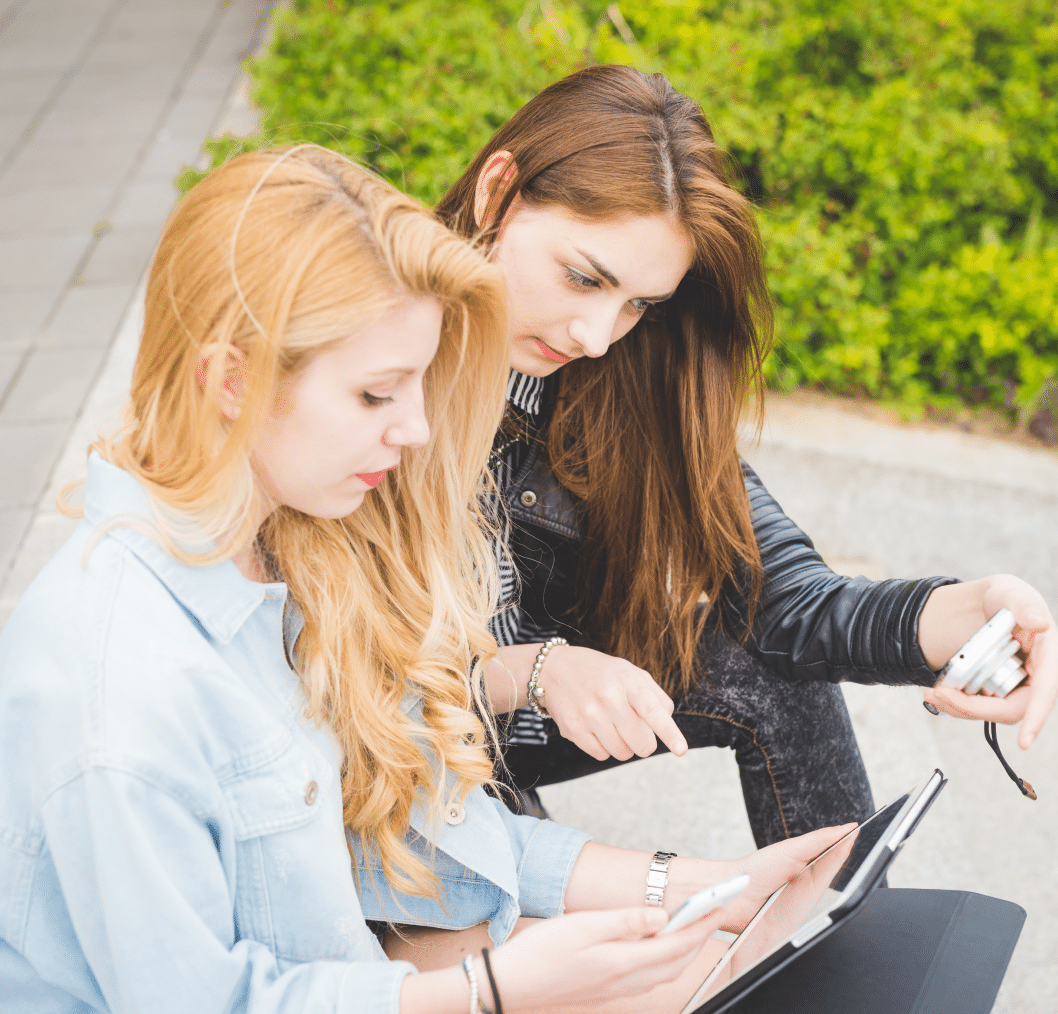 Two young women engaged in discussion while using a tablet and smartphone, emphasizing social media interaction and connectivity in a casual outdoor setting.